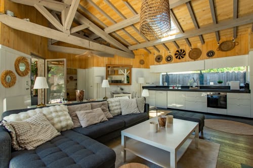 Intérieur de villa contemporaine en open space avec canapé sectionnel noir, cuisine blanche, poutres en bois et paniers décoratifs à La Loge Du