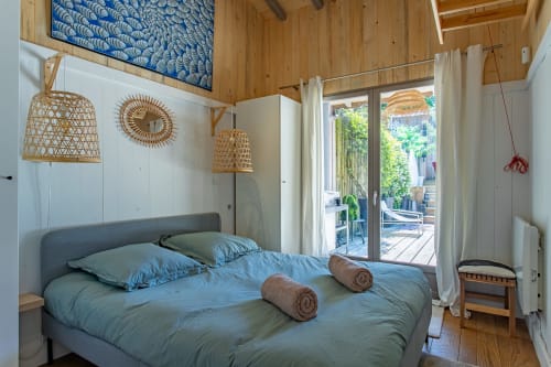 Chambre lumineuse à la villa La Loge Du Bassin avec literie bleue, lampes suspendues en rotin, plafond en bois et portes donnant sur le jardin