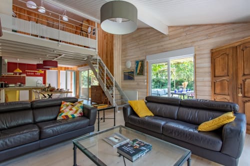 Salon moderne de villa avec canapés en cuir noir, accents jaunes, poutres en bois, mezzanine et vue sur le jardin à La Loge