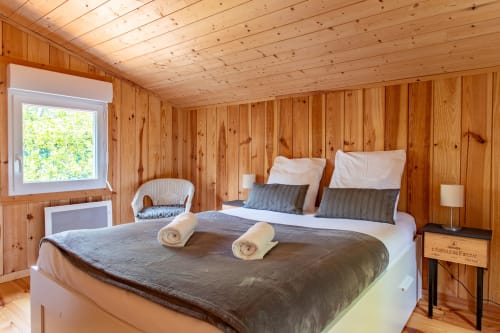 Villa chambre cosy en bois à La Loge Du Bassin : lit double, serviettes roulées, fauteuil en osier, vue jardin par la fenêtre