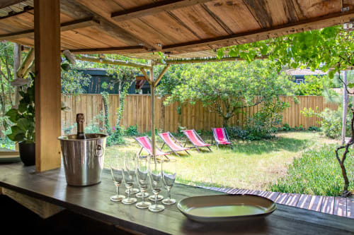 Vue sur le jardin depuis la terrasse couverte de la villa La Loge Du Bassin avec verres de vin, seau à glace, transats et verdure luxuriante