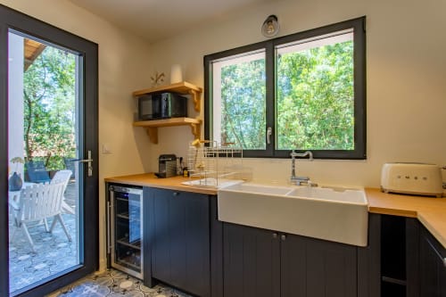 Kitchenette moderne avec évier, comptoirs en bois, armoires noires et fenêtres donnant sur la verdure à la villa La Loge Du Bassin