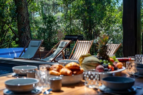 Espace repas extérieur à la villa La Loge Du Bassin avec transats rayés, piscine, fruits frais et jardin ombragé