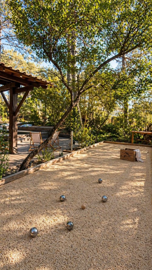 Jeu de boules en plein air sur la terrasse de graviers de la villa La Loge Du Bassin, ombragée par de grands arbres avec pergola en bois et piscine