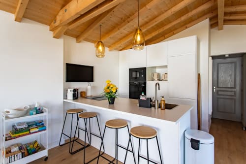 Cuisine blanche moderne avec poutres en bois, îlot de repas et luminaires jaunes à la villa La Palombière