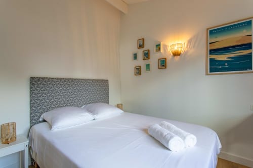 Chambre lumineuse avec tête de lit géométrique, literie blanche, serviettes roulées, gravures côtières encadrées et applique murale à La Loge Du