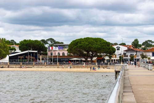 Vue du front de mer d'Andernos-les-Bains avec plage, jetée, rivage sableux et bâtiments en bord de plage sous un ciel nuageux