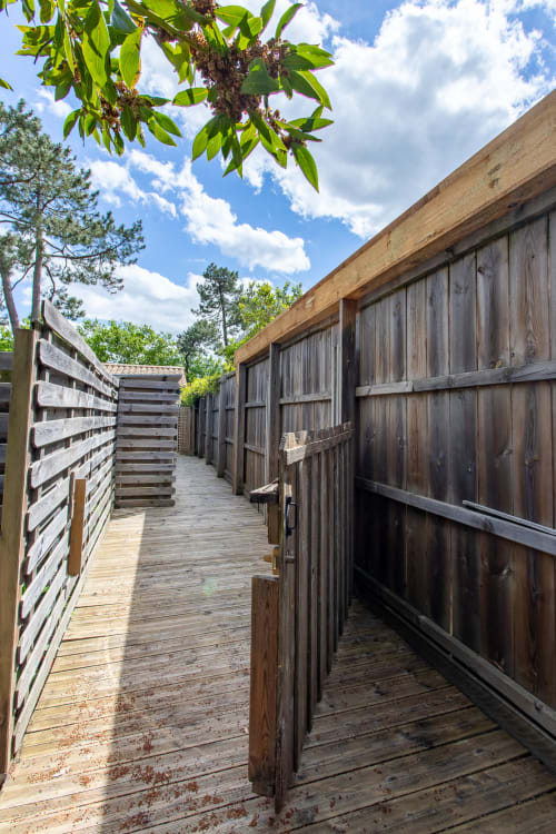 Passerelle en bois entre les écuries rustiques de la villa La Loge Du Bassin à Andernos-les-Bains, vue sur le jardin sous ciel bleu