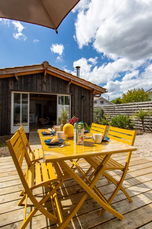 Ensemble table et chaises de salle à manger jaunes sur une terrasse en bois face à une villa en bois sombre, parasol et ciel bleu