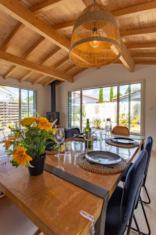 Salle à manger moderne de la villa La Palombière avec plafond à poutres apparentes, suspension en matière tressée, table dressée aux fleurs jaunes
