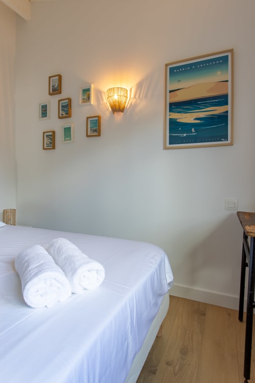 Chambre moderne à la villa La Loge Du Bassin avec lit blanc, serviettes roulées, applique murale en fibres tressées et œuvres côtières