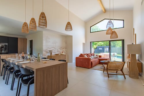 Intérieur de villa moderne avec luminaires en suspension tressés, îlot de salle à manger en bois, canapé corail et vue sur la piscine à La Parenthèse