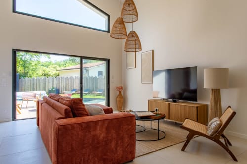 Salon de villa moderne avec canapé rouille, meuble TV en bois, luminaires suspendus tissés et vue sur la piscine par portes vitrées