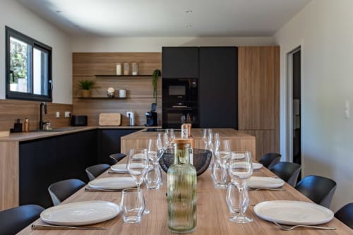 Salle à manger de villa contemporaine avec table en bois dressée pour le dîner, chaises noires, cuisine ouverte aux façades bois et noir à La