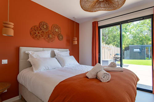 Chambre moderne avec mur d'accent orange vibrant, literie blanche, luminaire suspendu en osier et portes vitrées ouvrant sur piscine