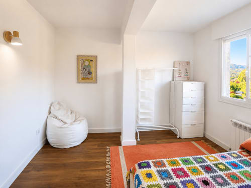 Dormitorio luminoso en la casita Nova Anduriña de Holibai, Gondomar, con muebles blancos, puf, alfombra colorida y vista al jardín