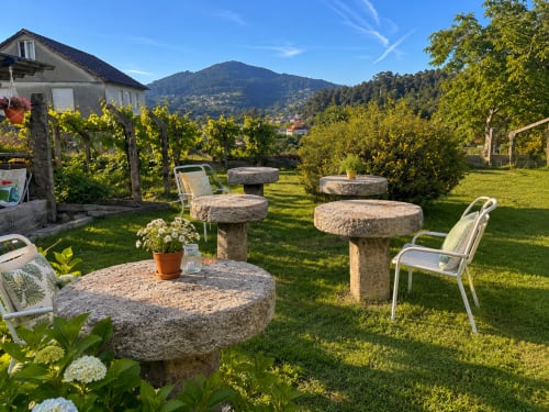 Sereno jardín de la casita Holibai en Gondomar con mesas de piedra, sillas metálicas, plantas floridas y vistas a la montaña.