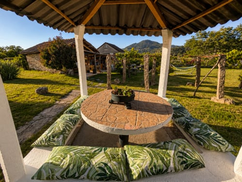 Pérgola cubierta con mesa de piedra redonda y asientos con cojines verdes con vistas al jardín de la casa Holibai en Gondomar