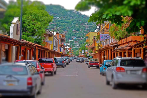 Rue coloniale colorée à Zihuatanejo avec voitures garées, bâtiments orangés et paysage verdoyant en arrière-plan