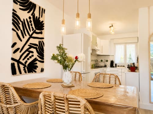 Salle à manger moderne avec table en bois, chaises tissées, œuvre d'art noir de feuilles, et luminaires suspendus dans l'appartement de L'Île-Rousse