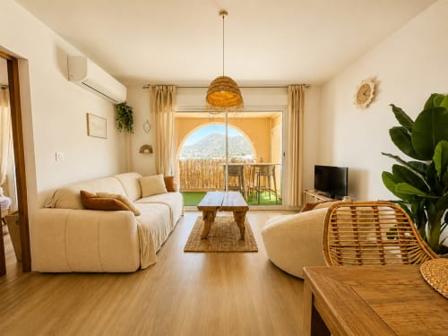 Lumineux séjour aux canapés courbes crème, mobilier bois et vue montagne depuis le balcon de l'appartement L'Île-Rousse