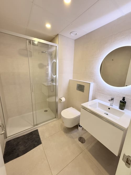 Baño moderno con ducha abierta, lavamanos flotante, espejo redondo y accesorios blancos en casita de Benalmádena