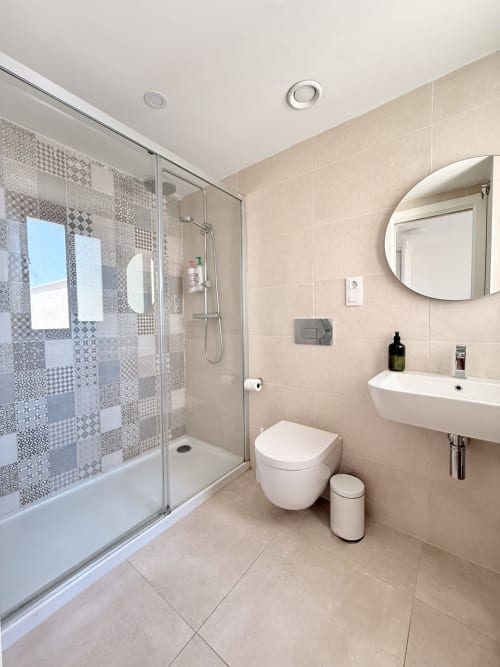 Baño moderno en casa de campo de Benalmádena con ducha abierta, lavamanos mural, inodoro, espejo redondo y azulejos beige