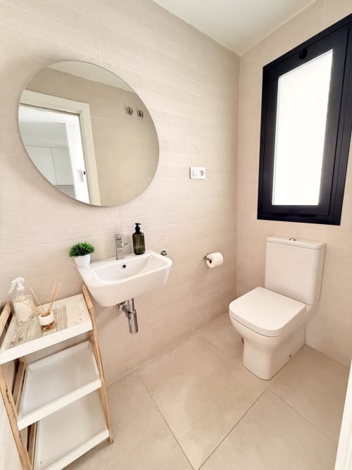Baño moderno con espejo redondo, lavabo blanco, inodoro y suelos de azulejo en casa rural de Benalmádena