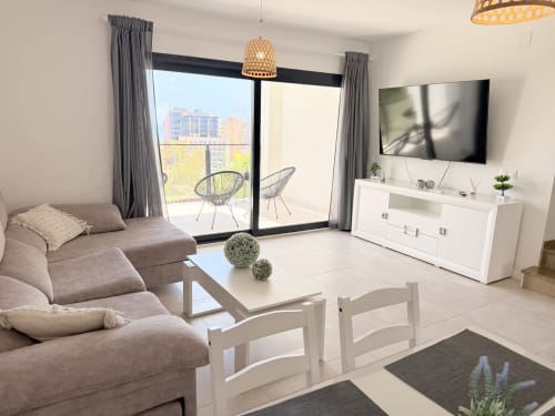 Luminoso salón moderno en casa de Benalmádena con sofá gris, muebles blancos, TV de pantalla plana y vistas de la ciudad desde la terraza