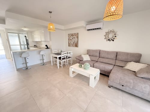 Sala de estar de planta abierta moderna con sofá gris, mesa comedor blanca, cocina con barra y lámparas colgantes tejidas