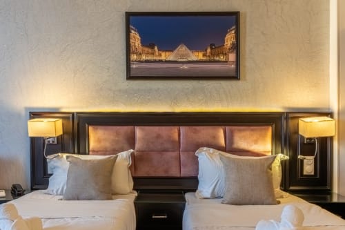 Deux lits jumeaux dans une chambre de l'Hôtel Royal Paris Ivry avec tête de lit, lampes de lecture et artwork de la pyramide du Louvre au-dessus