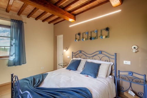 Spazios camera da letto all'Agriturismo Marinello di Pienza con letto in ferro blu, biancheria bianca, travi in legno a vista e vista sulla città