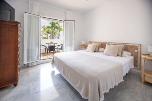Dormitorio luminoso frente al mar en BH Marbella con cama de ratán, ropa blanca, vistas al mar y palmeras a través de terraza
