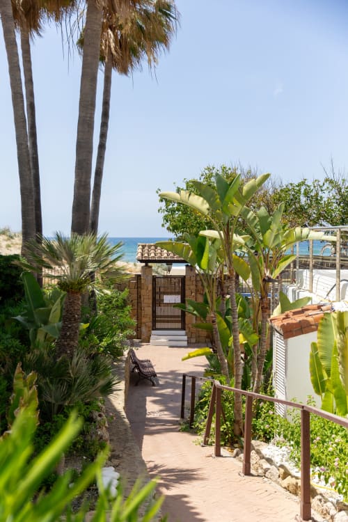 Sendero tropical frente al mar en cabaña Maui, BH Marbella, con palmeras, platanales y vistas al mar a través de arco de piedra