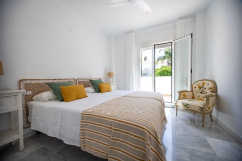 Dormitorio luminoso en la cabaña BH Marbella Maui con cama de ratán, puertas con vista al océano, manta a rayas y butaca