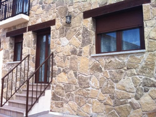 Entrada de fachada de piedra de Alojamientos Botica Rural con puerta de madera, ventana y barandilla metálica en La Cabrera
