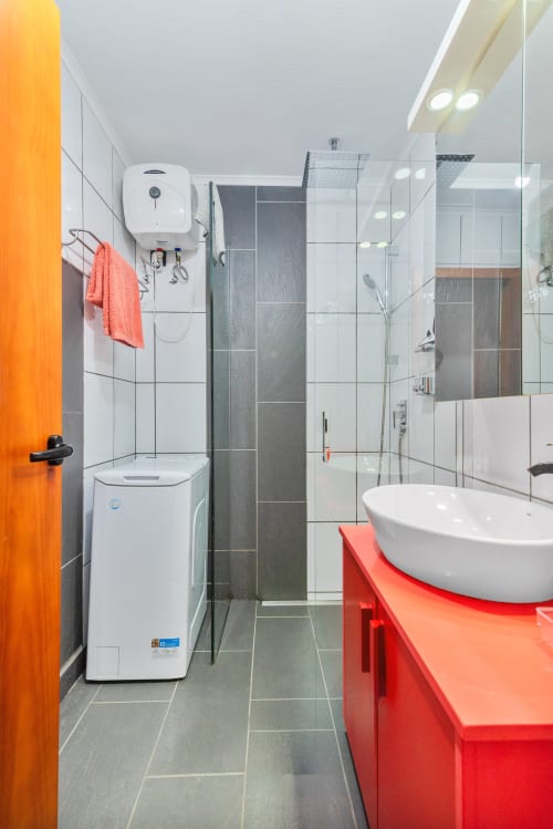 Baño moderno con azulejos blancos, lavamanos rojo, mampara de ducha de vidrio y puerta naranja en Home Stay Gran Canaria