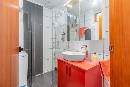 Baño moderno con tocador rojo, lavamanos blanco, cabina de ducha de cristal y detalles naranjas en Home Stay Gran Canaria