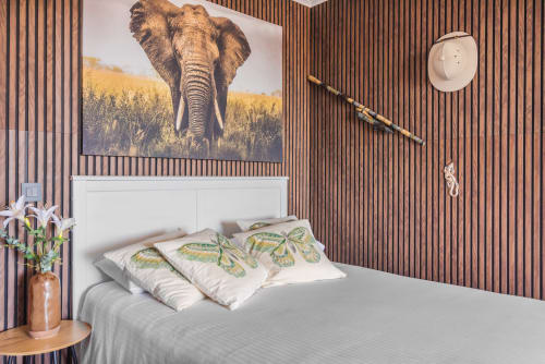 Dormitorio moderno con cuadro de elefante sobre cama blanca, almohadas con mariposas, pared de madera, flores en maceta y vista al mar