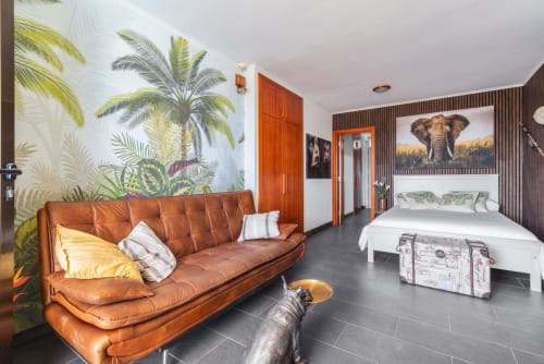 Moderno estudio con sofá de cuero marrón, papel tapiz tropical de palmeras, puertas de madera roja y cama con arte de elefantes