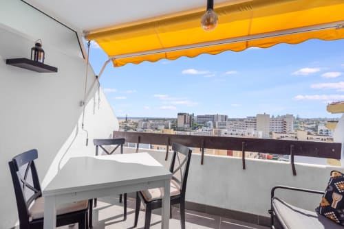 Terraza soleada del apartamento Las Arenas en Gran Canaria con toldo amarillo, mesa de comedor, vistas a la ciudad y al mar