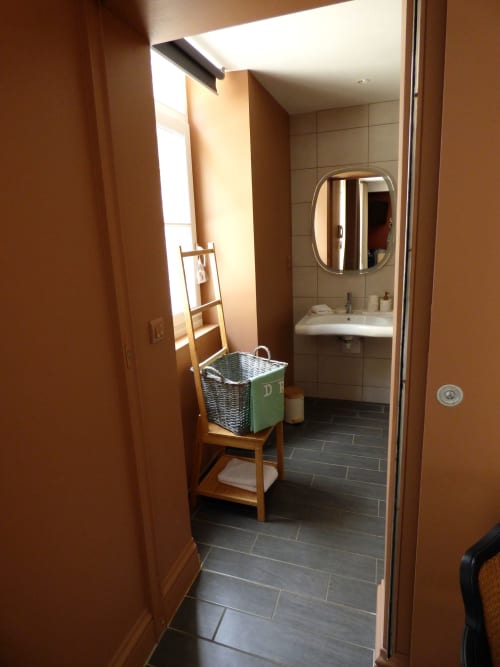 Bathroom entrance at Les Rives De La Tronne, Mer - sink, round mirror, dark tile flooring, laundry basket, and bright natural