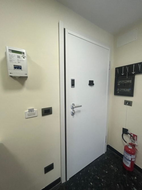 Ingresso appartamento Luan House a Genova con porta bianca, contatore elettrico, termostato e estintore su piastrelle scure