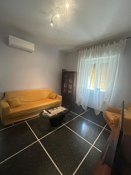 Luminosa living room di Luan House Genova con divano giallo, pavimento in piastrelle nere e tende trasparenti con vista sulla città