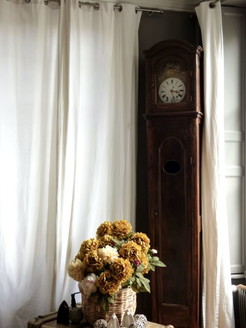 Elegant corner with golden flowers in basket, antique wooden grandfather clock, and cream curtains at Les Rives De La Tronne