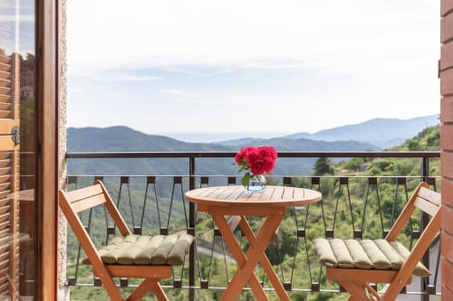 Balcon de l'hôtel Allegria à Bajardo avec deux chaises en bois, petite table avec fleurs rouges et vue sur les montagnes