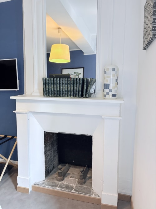 Élégante cheminée avec manteau blanc, lampe suspendue jaune et mur d'accent bleu foncé dans la Suite Camille Claudel à Au Richebourg