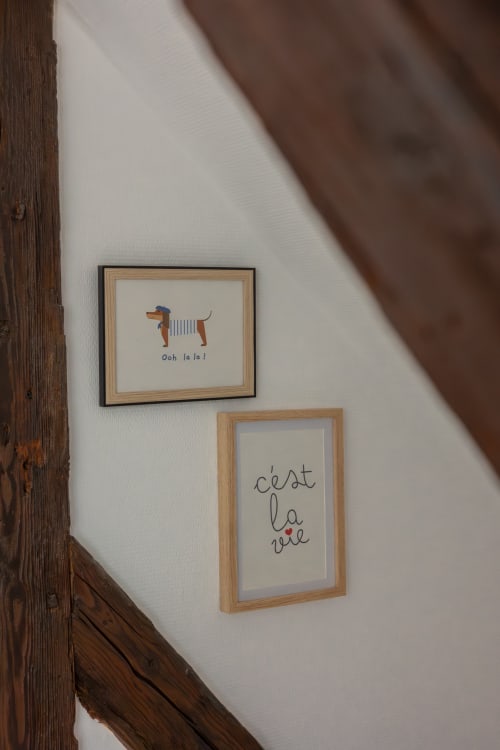 Two framed artworks on white wall at Le JO·LIE Duplex: dachshund with 'Ooh la la!' and French text 'c'est la vie'
