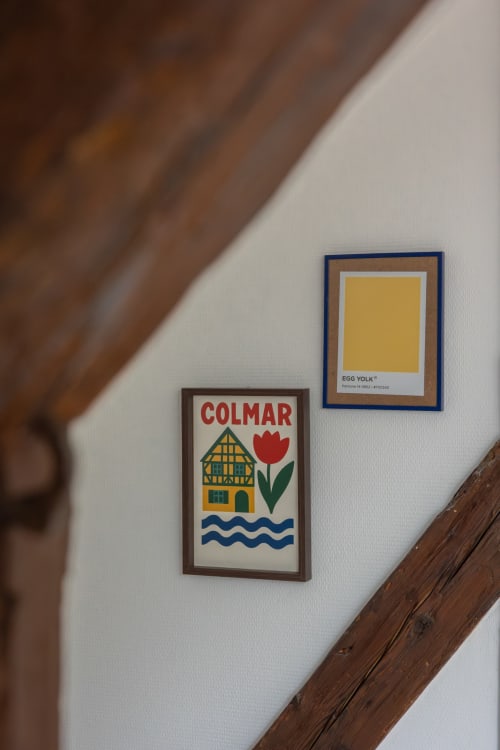 Two framed artworks on white wall: Colmar postcard with yellow house and red tulip, Egg Yolk color swatch. Wooden beam below.