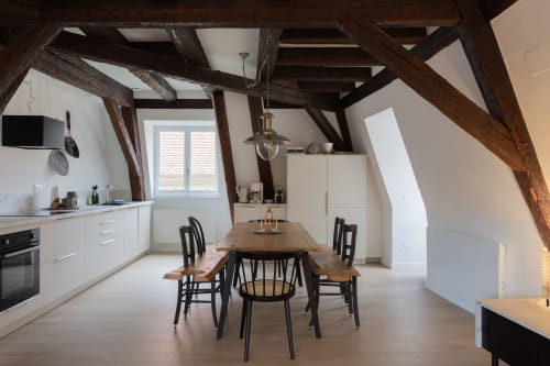 Modern loft kitchen and dining area with exposed wooden beams, white cabinetry, wooden table, and black chairs in Colmar